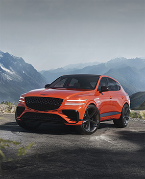 An orange-hued Genesis GV80 Coupe Concept vehicle is parked on a road with rugged mountains in the background. The headlights are on, and the front wheels are slightly turned with skid marks visible.