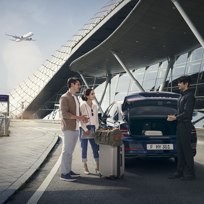 공항 앞에서 여행객이 차량 픽업 서비스를 받는 장면입니다. 열린 트렁크 앞에서 짐을 싣는 모습이며, 직원이 고객을 맞이하고 있습니다. 배경에는 공항과 비행기가 보이며, 에어포트 서비스 분위기를 연출합니다.