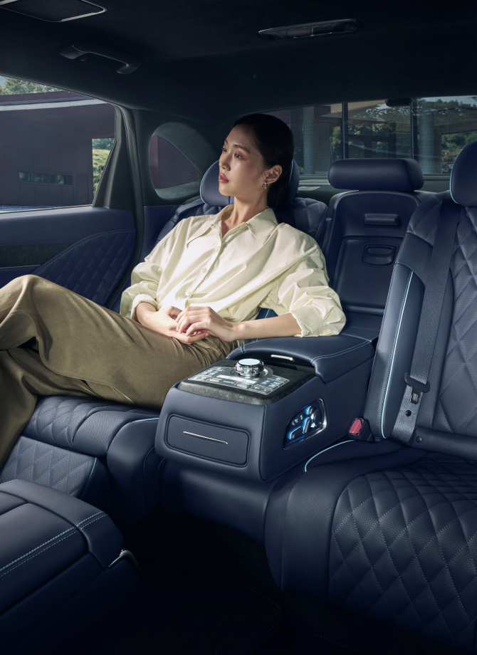 A young woman is shown sitting in the back of a car, resting her arm on the centrally placed armrest. The view is seen from the front seat, giving the impression of a spacious back seat.